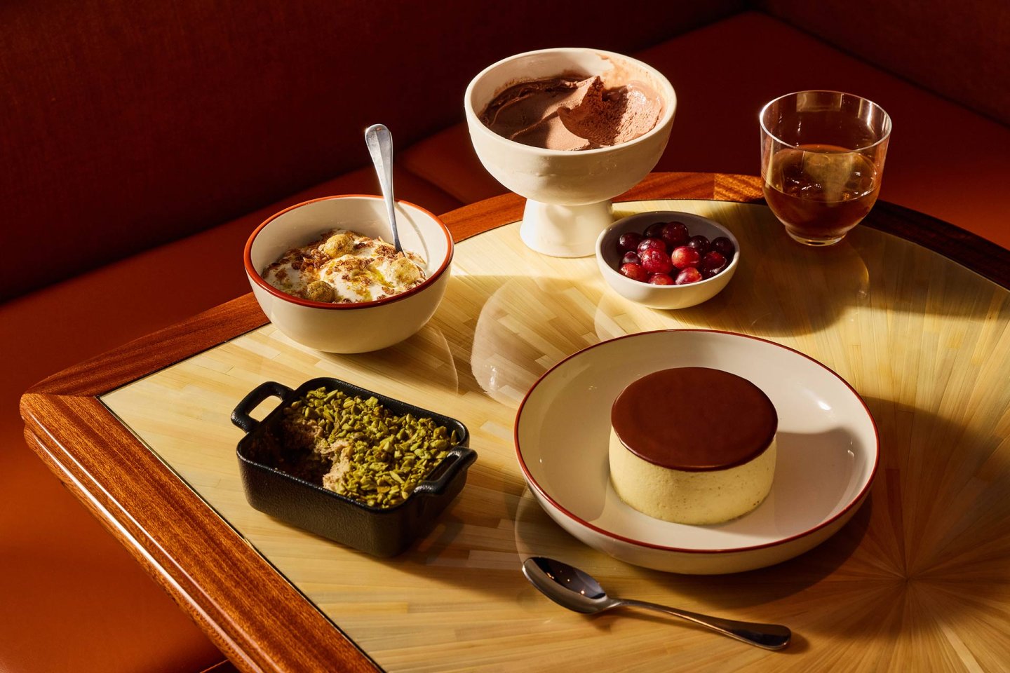 A table set with five desserts: a bowl of chocolate mousse, a glass of iced drink, a dish of red grapes, a baked dessert in a small black dish, a bowl of baked pudding, and a round cheesecake with a chocolate top.
