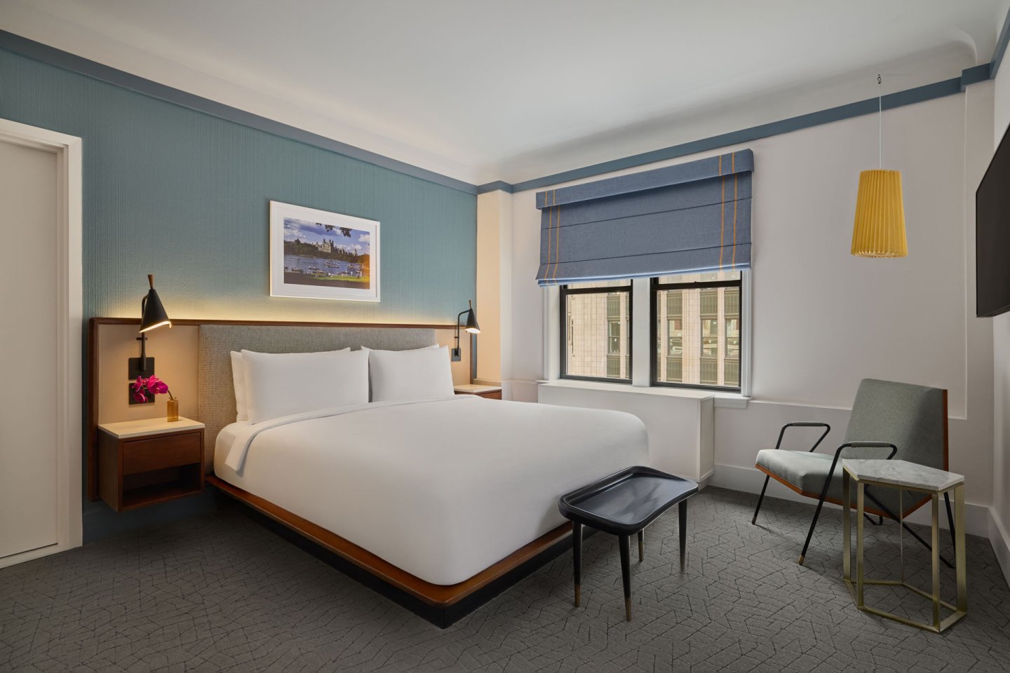 Modern hotel room with a king bed, two nightstands, lamps, wall art, a window with blue blinds, a chair, a small table, and a bench at the foot of the bed. The decor is minimalist and inviting.