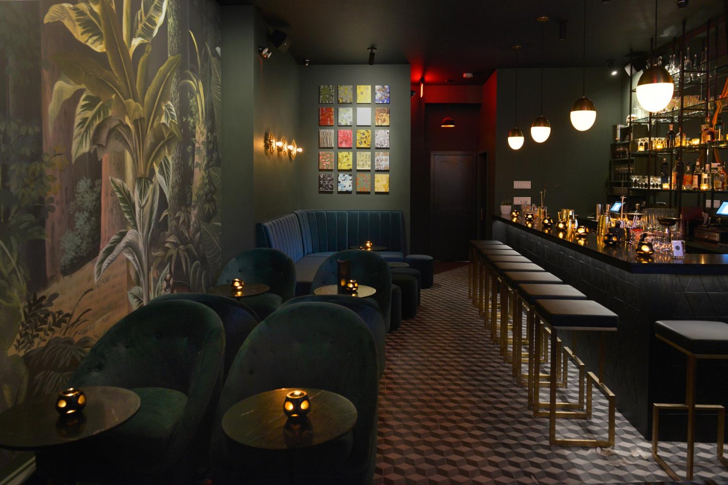 A dimly lit, elegant bar with velvet seating, a long counter with high stools, ornate wall art, and pendant lights. The floor has a geometric pattern, and the atmosphere is cozy and sophisticated.