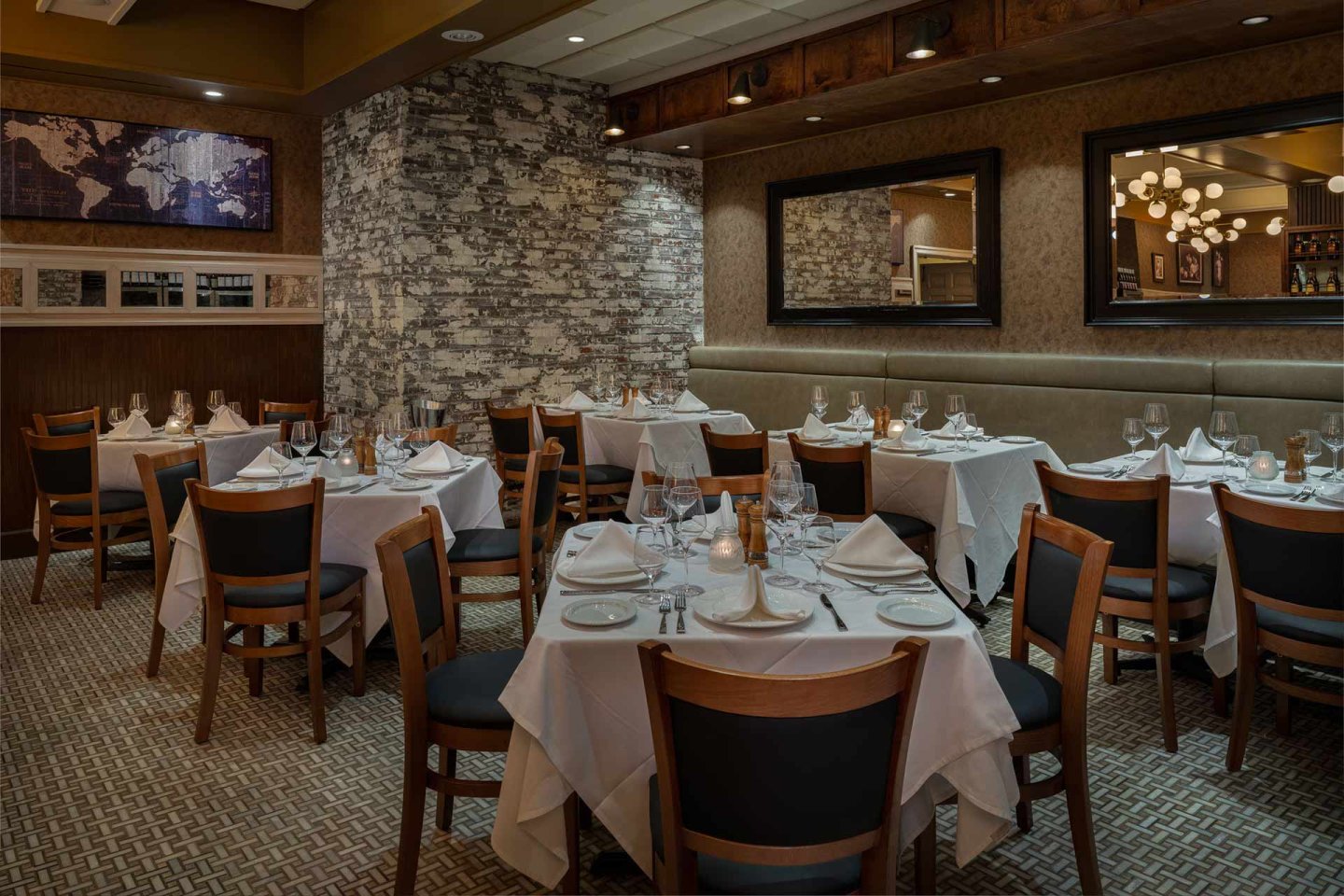 Elegant restaurant dining room with neatly set tables covered in white tablecloths, wooden chairs, exposed stone accent wall, warm lighting, mirrors, and a map on the wall, creating an inviting and upscale atmosphere.