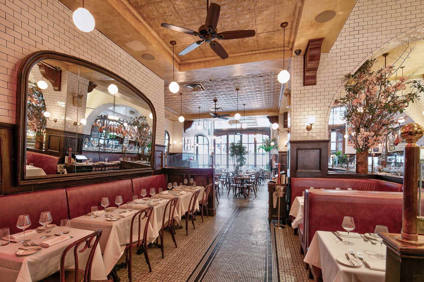A stylish restaurant with white tiled walls, red upholstered booths, wooden chairs, tables covered in white cloths, large mirrors, globe lights, and indoor plants, creating a warm and inviting atmosphere.