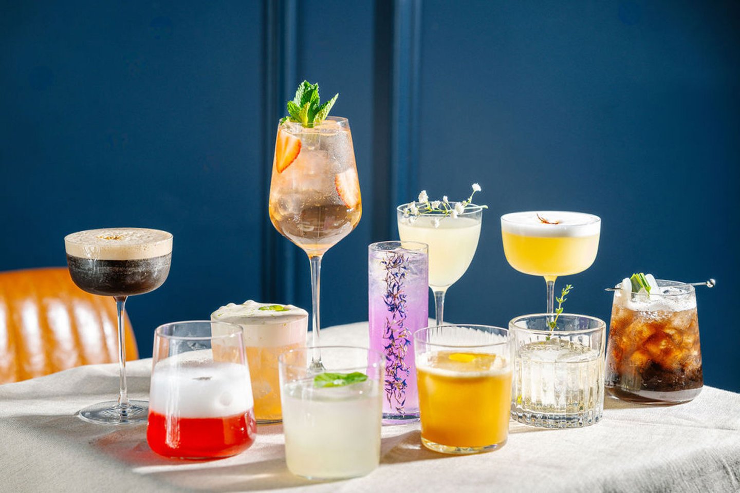 A variety of colorful cocktails displayed on a table, set against a dark blue background. There are ten drinks in different types of glasses, some with garnishes like mint leaves, lemon slices, and ice cubes. Each drink varies in color and presentation.