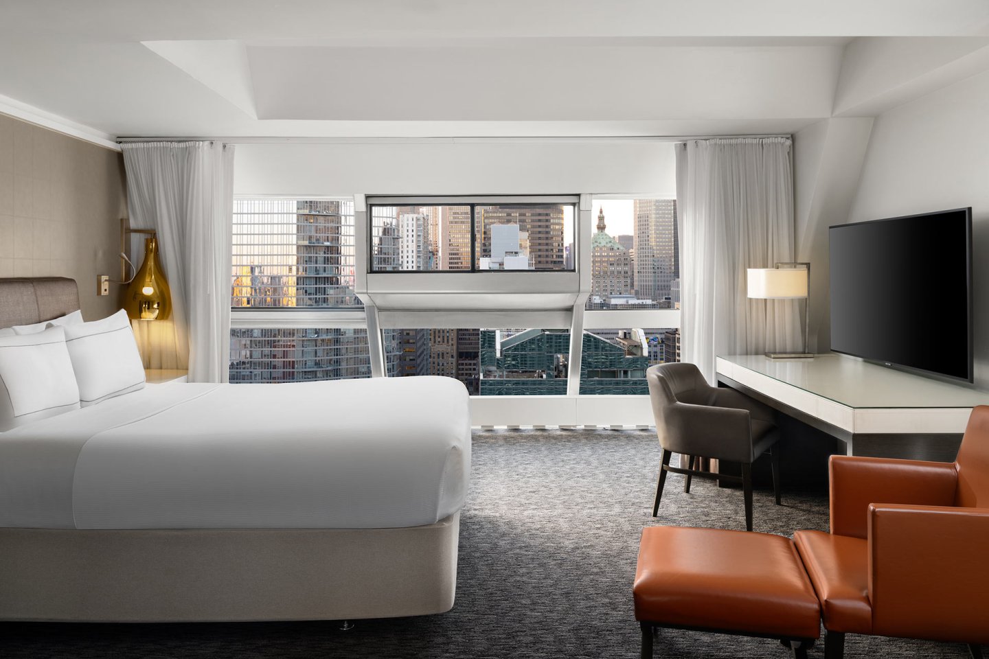 A modern hotel room with a large bed, a desk with a chair, a TV, and an orange armchair with an ottoman. Large windows offer a city skyline view with tall buildings in the background.