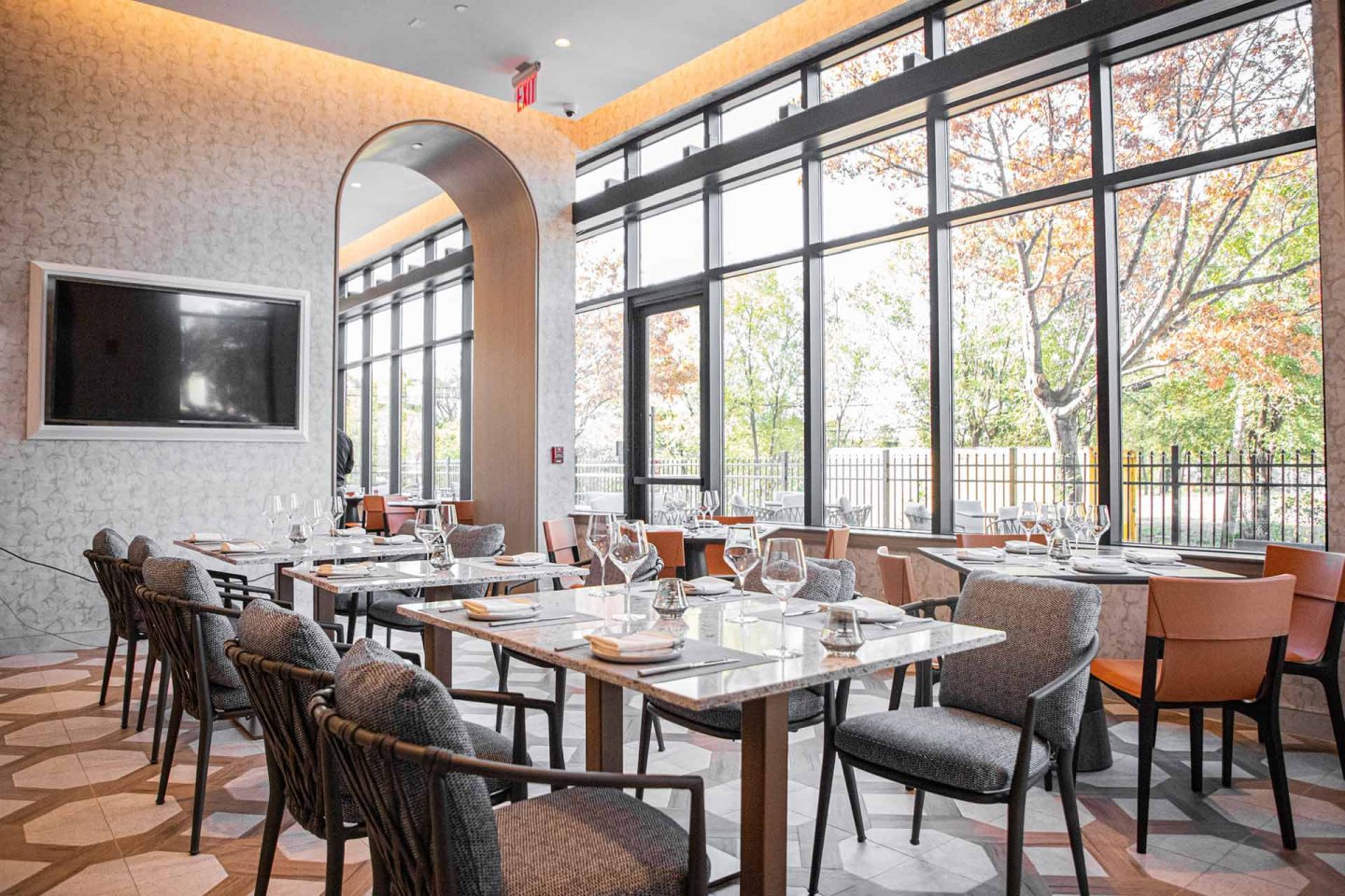 Bright, modern restaurant interior with large windows, marble tables set with glassware, cushioned chairs, and a wall-mounted TV. Natural light fills the space, highlighting trees visible through the windows.