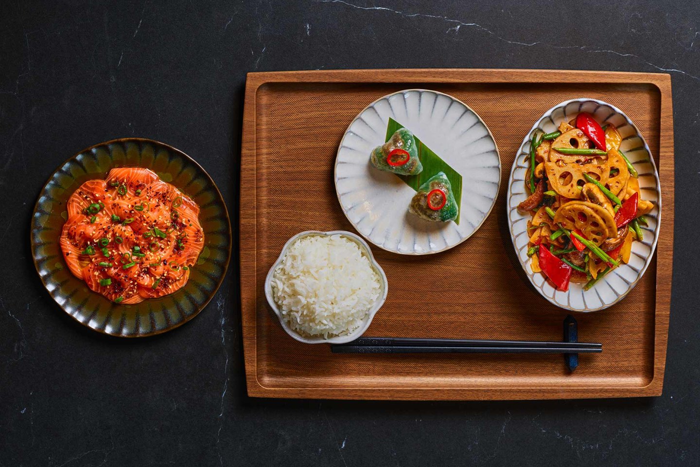 A wooden tray holds a bowl of white rice with chopsticks, a plate of sliced lotus root with peppers, a small dish with a wrapped bite, and a separate plate of sliced salmon topped with herbs. All dishes are neatly arranged.