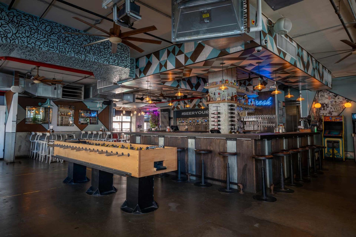 Spacious modern bar with geometric decor, a large square bar counter surrounded by stools, colorful lighting, and a foosball table in the center. The space has industrial elements and artistic wall designs.
