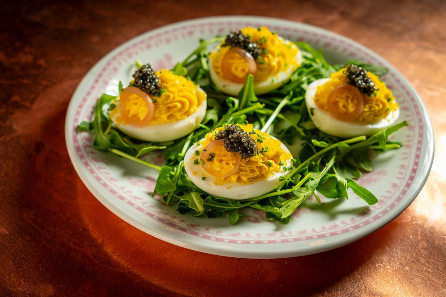 Four deviled eggs topped with caviar and herbs are arranged on a bed of arugula, served on a white plate with a pink decorative rim, sitting on a copper-colored surface.