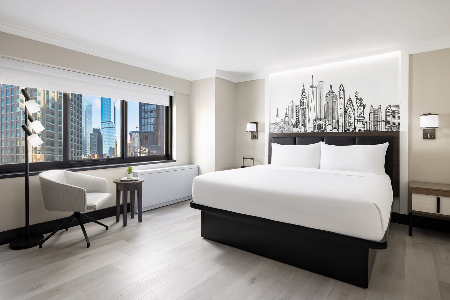 Modern hotel room with a large bed, cityscape artwork on the headboard, a white chair by a small table, a floor lamp, and large windows showing tall buildings outside.
