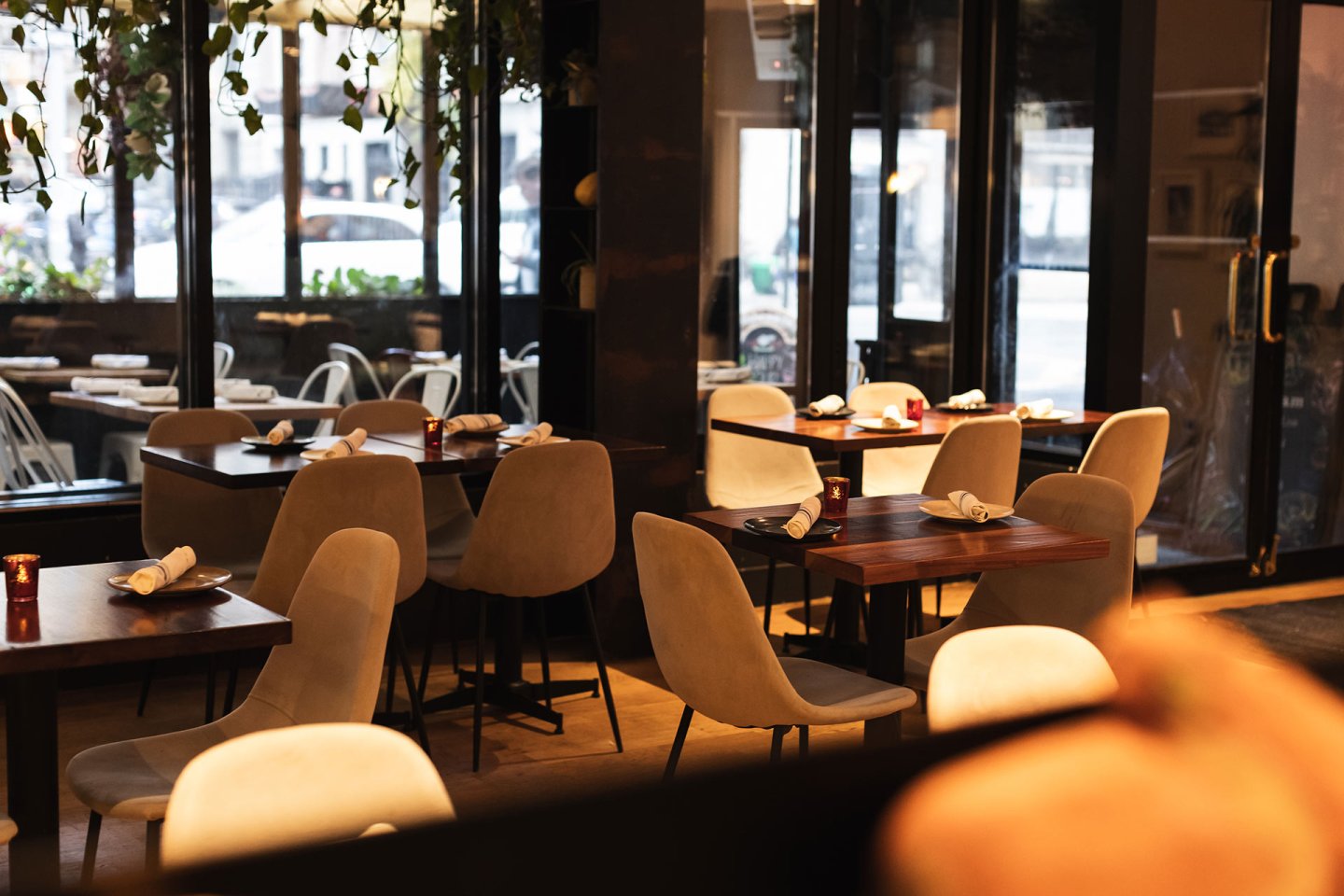 Modern restaurant interior with empty tables and beige chairs, neatly set with napkins and glasses. Warm lighting and large windows create a cozy, inviting atmosphere.