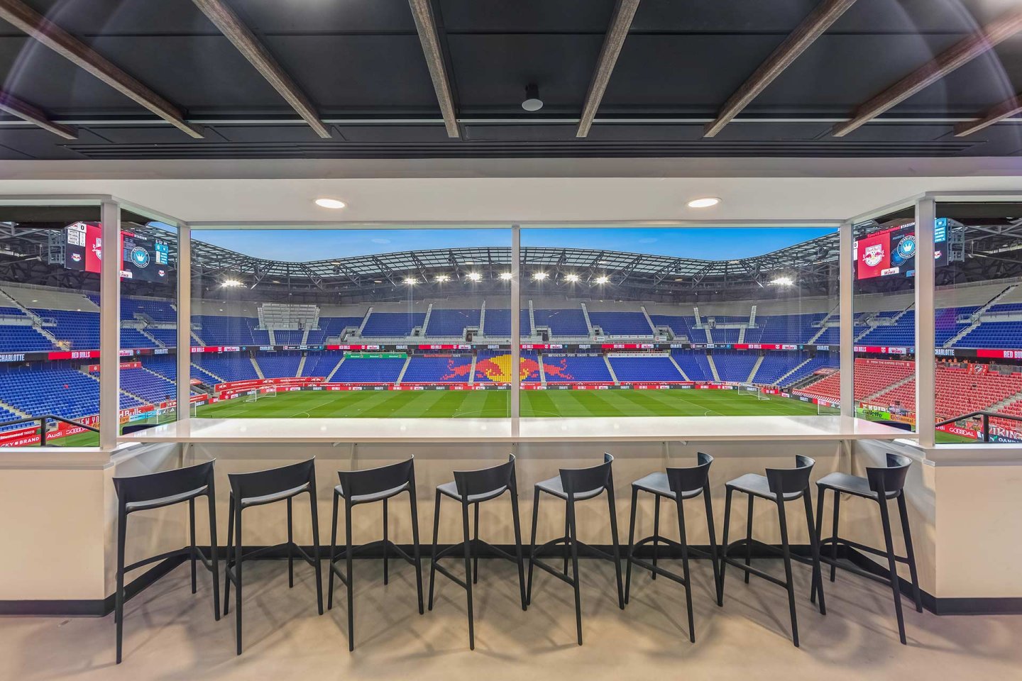 A row of black chairs lines a counter by large windows overlooking an empty soccer stadium with blue and red seats and a field featuring a yellow and red logo at the center.