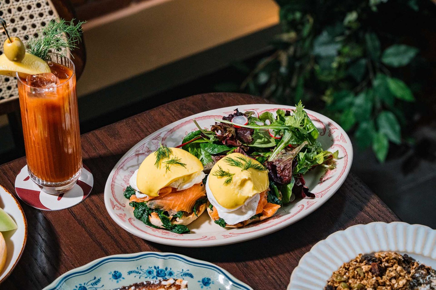 Plate with two eggs Benedict on smoked salmon and spinach, topped with hollandaise sauce and dill, served with a mixed green salad. A Bloody Mary cocktail sits nearby on the wooden table.