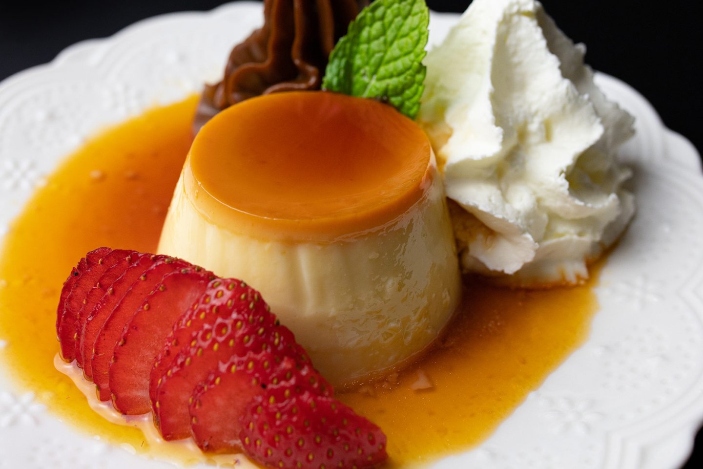A close-up of a flan dessert with caramel sauce, garnished with sliced strawberries, a dollop of whipped cream, a chocolate decoration, and a sprig of fresh mint on a white plate.