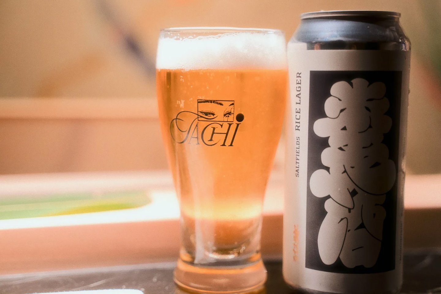 A can of Sapporo rice lager with bold black-and-white Japanese lettering sits next to a filled glass of beer on a table. The glass has "SACHI" and an eye logo printed on it.