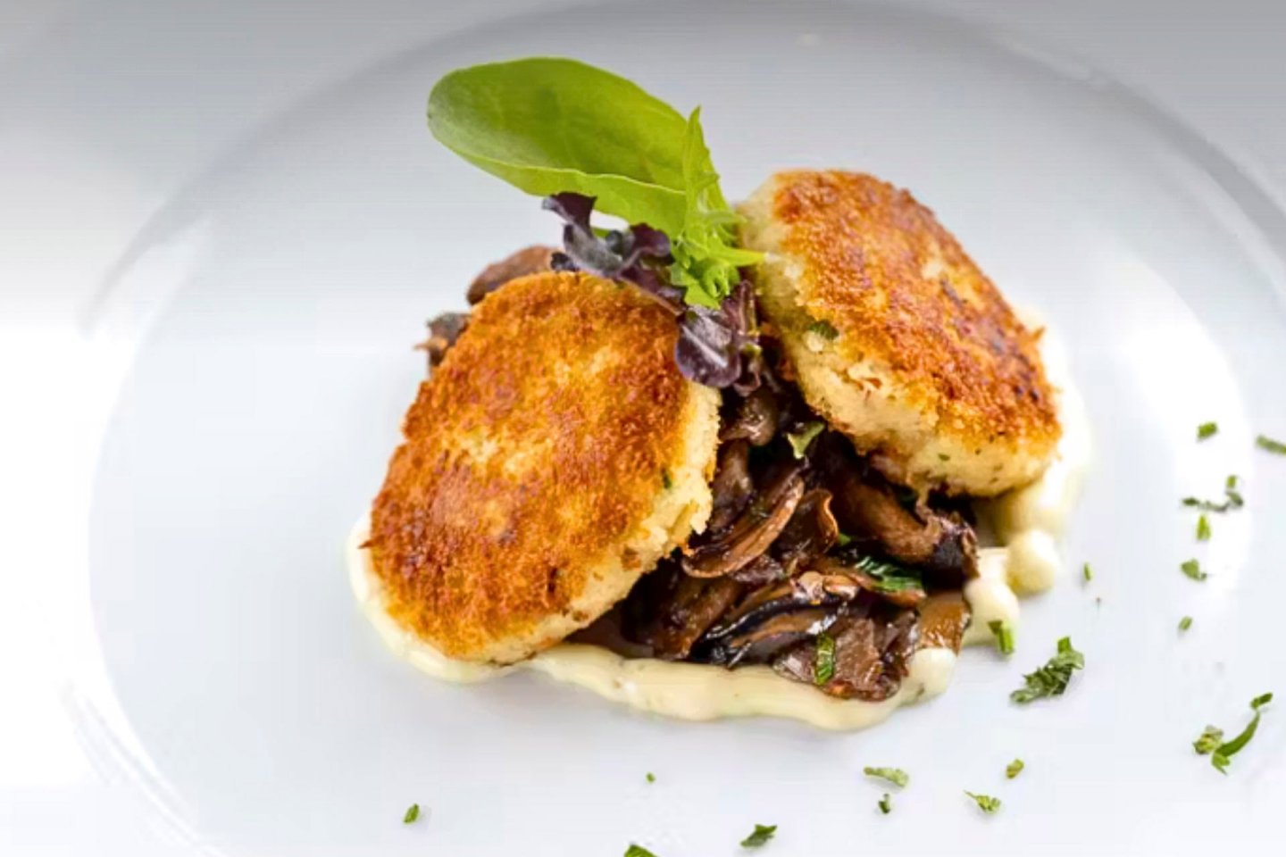 Two golden-brown crab cakes sit atop sautéed mushrooms, garnished with a leafy green and herbs on a white plate. A creamy sauce partially encircles the dish, adding a refined touch.
