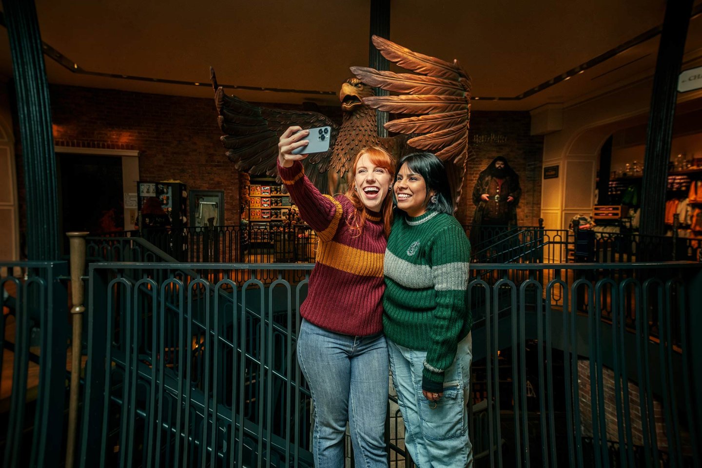 Two women wearing Hogwarts-themed sweaters take a selfie together in front of a large eagle statue inside a Harry Potter-themed shop or attraction, smiling and standing on a stairway.
