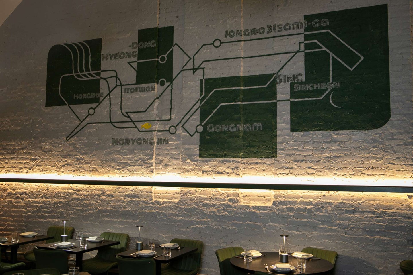 A painted subway map with Korean station names decorates a brick wall above a row of tables and green chairs in a modern, dimly lit restaurant.