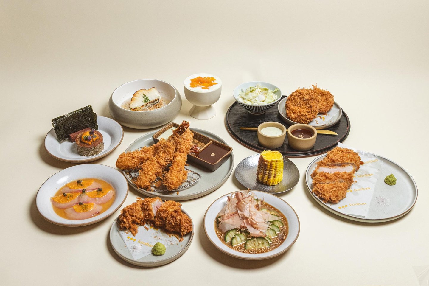 An assortment of Japanese dishes on plates, including fried cutlets, shrimp tempura, rice bowls, pickled vegetables, corn, dipping sauces, sushi, and dessert, all arranged neatly on a light background.