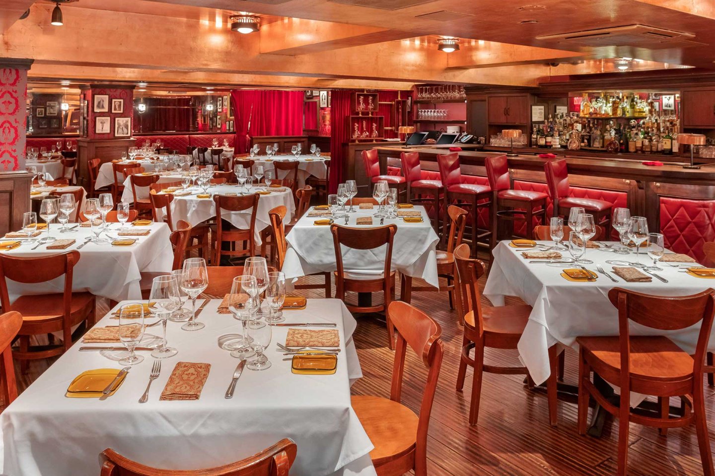Elegant restaurant with wooden chairs and white tablecloths, set for dining with glassware and plates. Red leather barstools line the bar area, and warm lighting creates a cozy, inviting atmosphere.