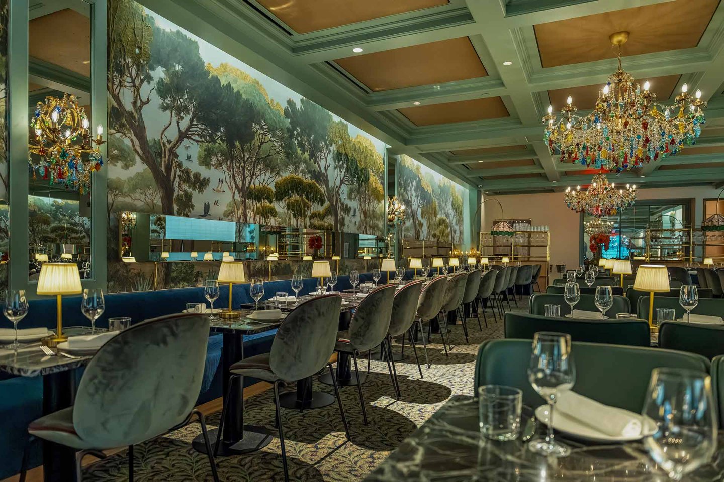 Elegant restaurant interior with chandeliers, velvet chairs, marble tables set for dining, and a mural of trees along the wall, creating a luxurious and inviting atmosphere.
