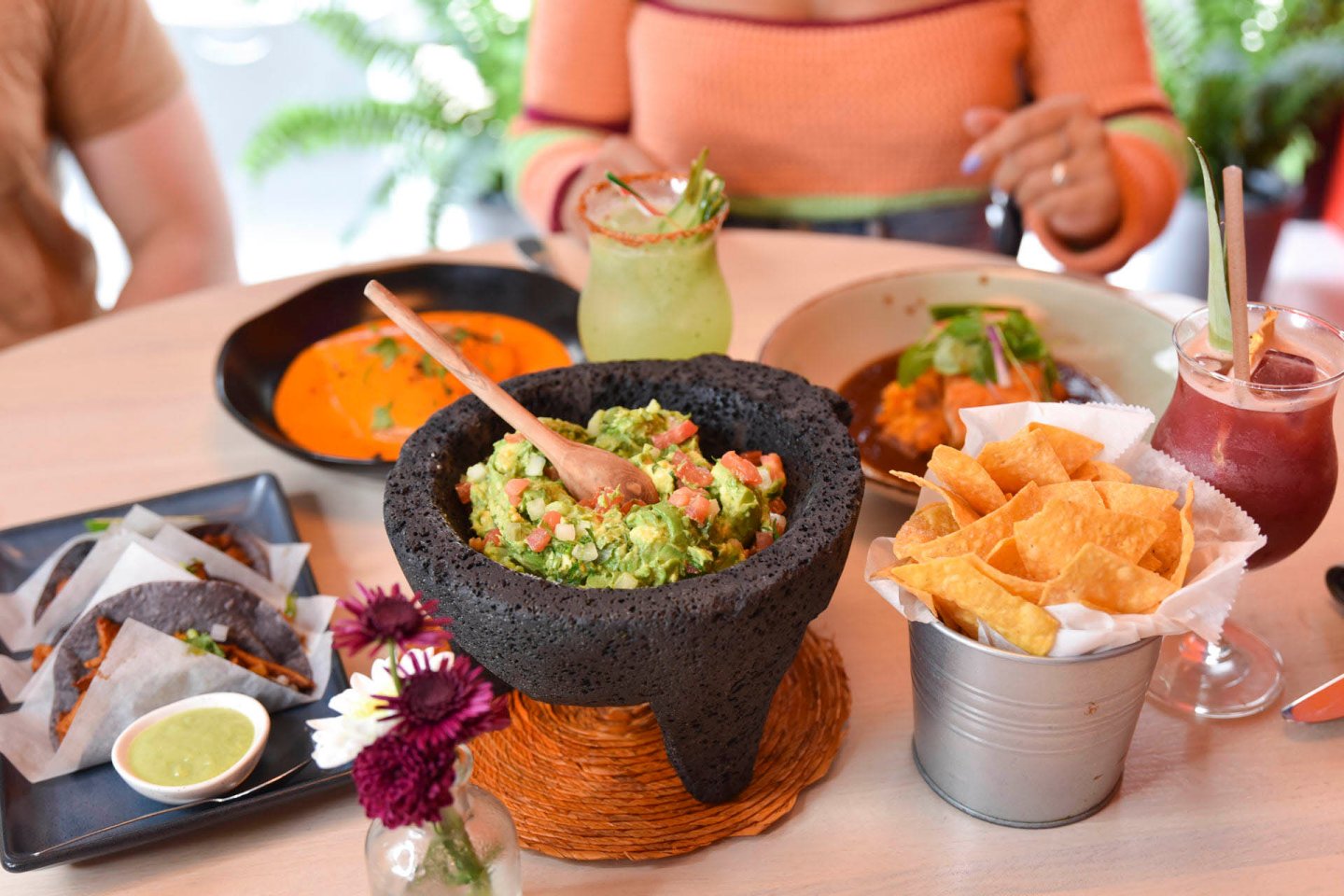A table set with various dishes: a large stone bowl of guacamole with a spoon, nachos in a metal container, a green drink, a red drink, black tacos with green sauce, and a dish with sauce. A person is in the background. Flowers nearby.
