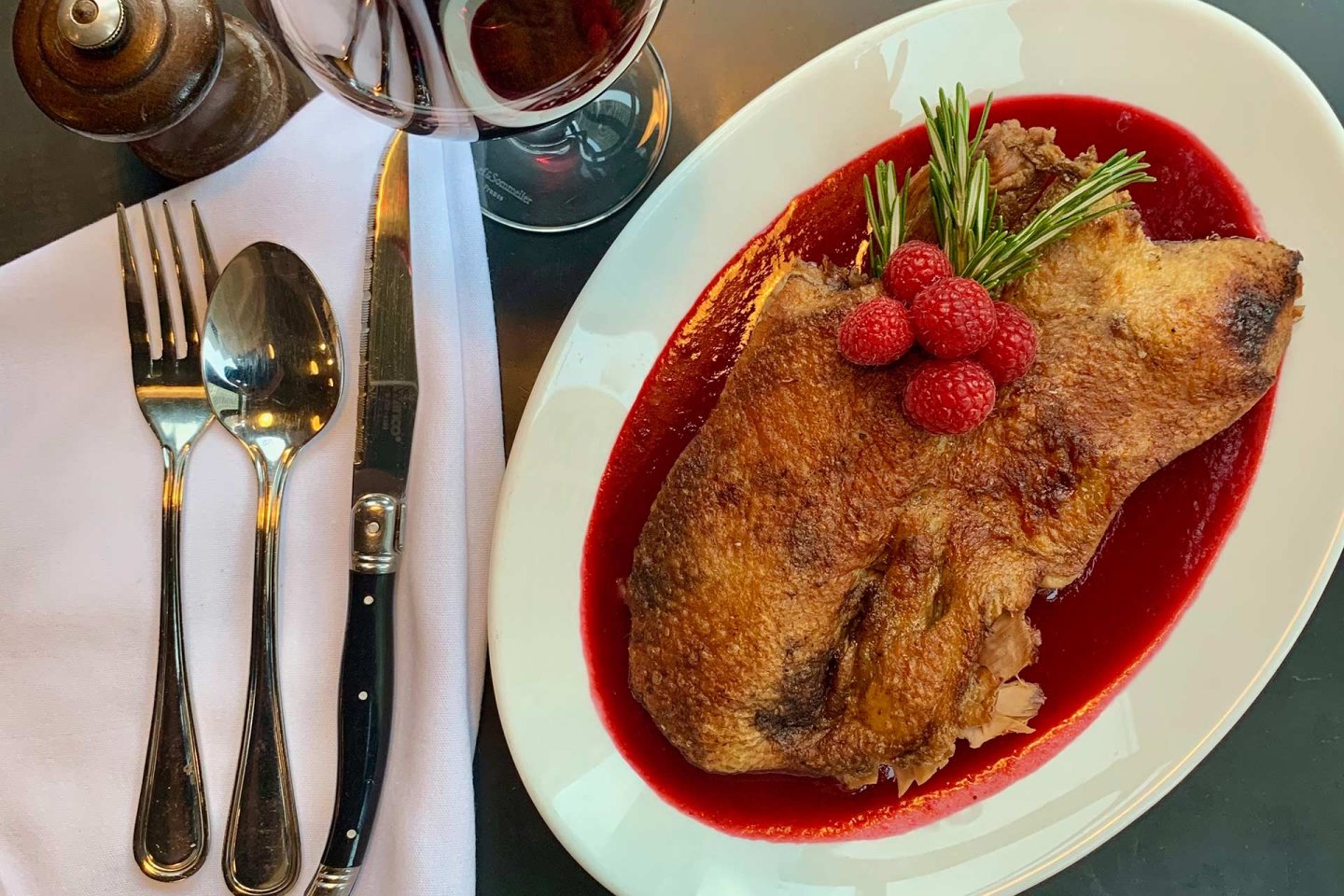 A plate with crispy roasted duck topped with raspberries and rosemary, served on a vibrant red sauce. Beside it are a fork, spoon, knife, and a glass of red wine on a white napkin.
