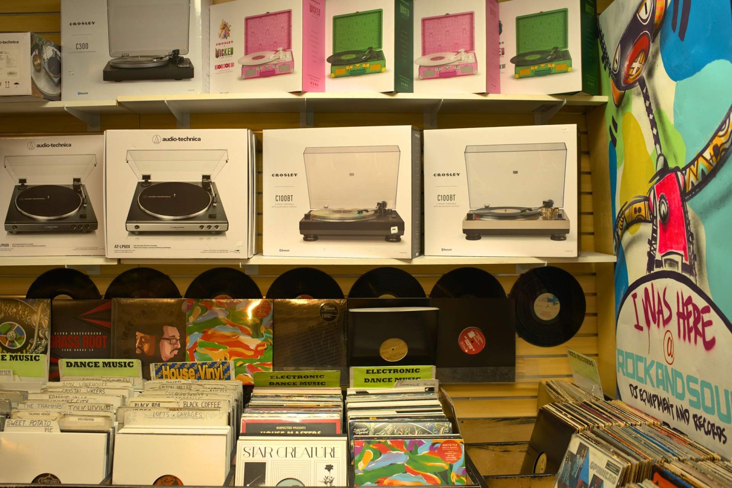 A display of record players and vinyl records in a music store. Turntables are stacked in boxes on shelves, and colorful album covers are arranged below. A mural with bold text is painted on the right side of the wall.