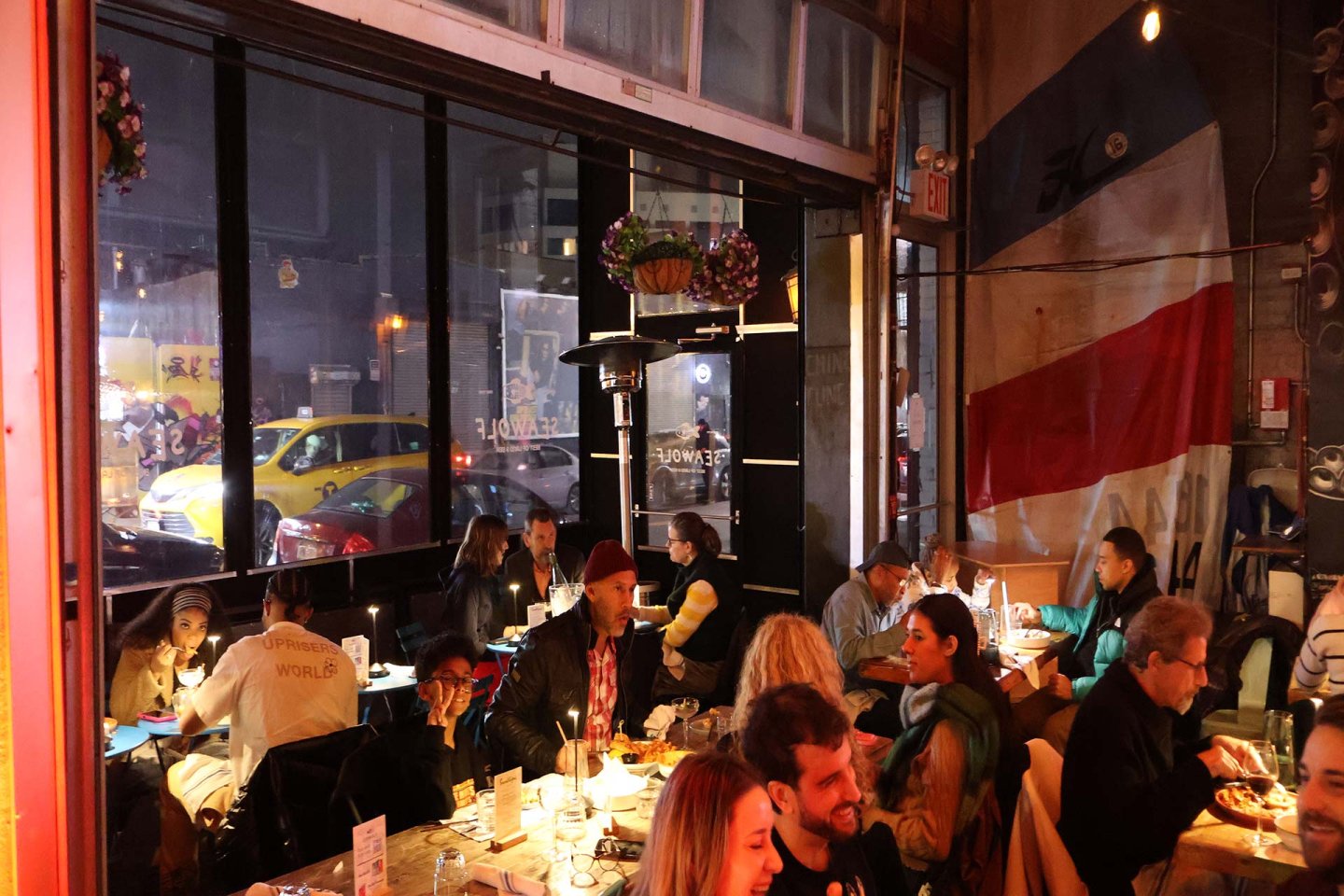 A busy, warmly lit restaurant with people sitting at tables eating and talking. Large windows show cars and city lights outside. An oversized flag hangs on the wall, adding to the lively atmosphere inside.