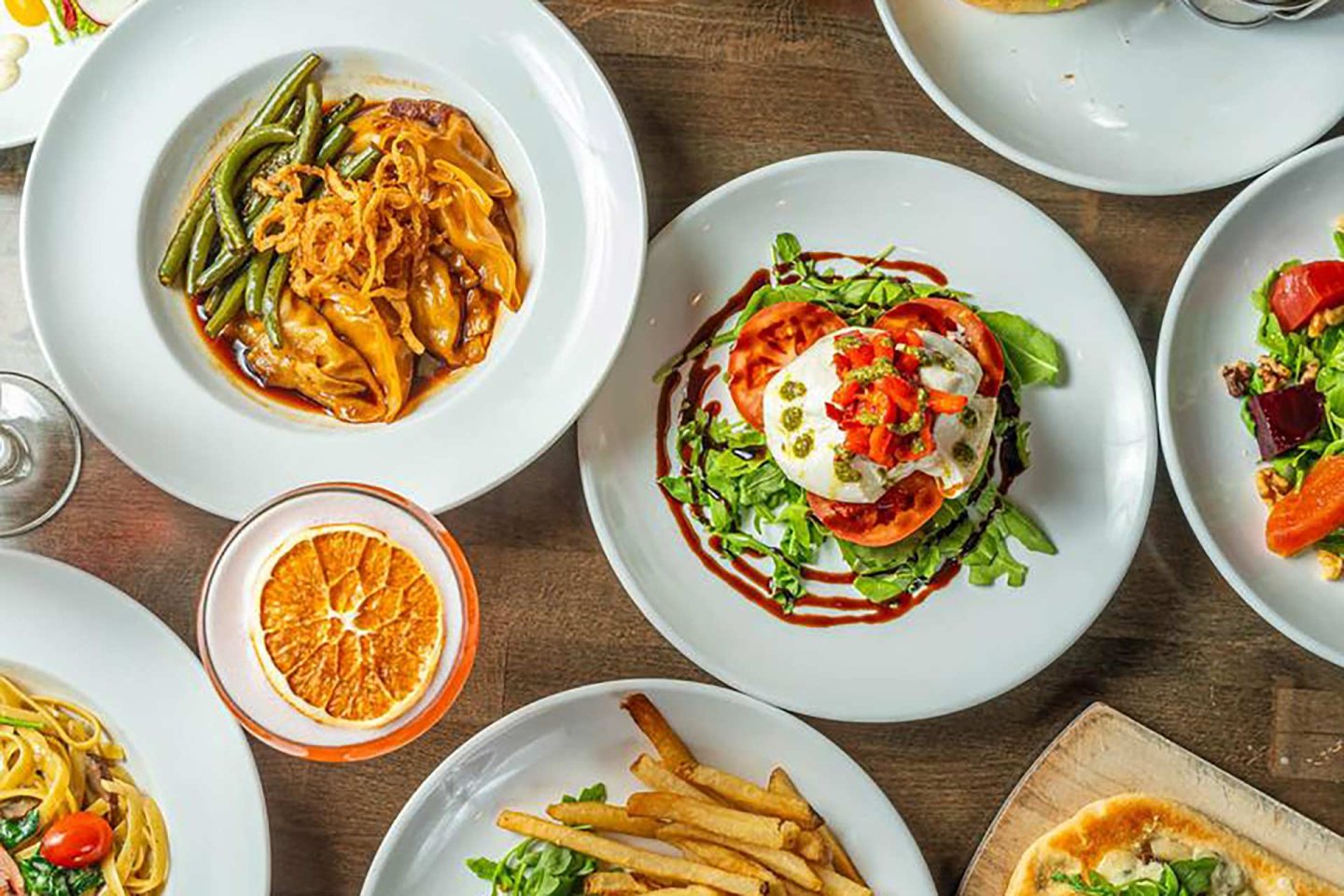 A table set with various dishes, including pasta, a salad with tomatoes and mozzarella, French fries, a meat dish with green beans, and a drink garnished with a slice of orange.