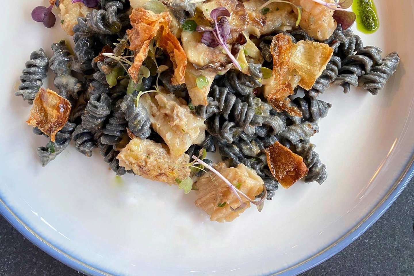 A close-up of a plate of dark-colored spiral pasta, topped with crispy golden pieces, microgreens, and a creamy sauce, served on a white dish with a blue rim.