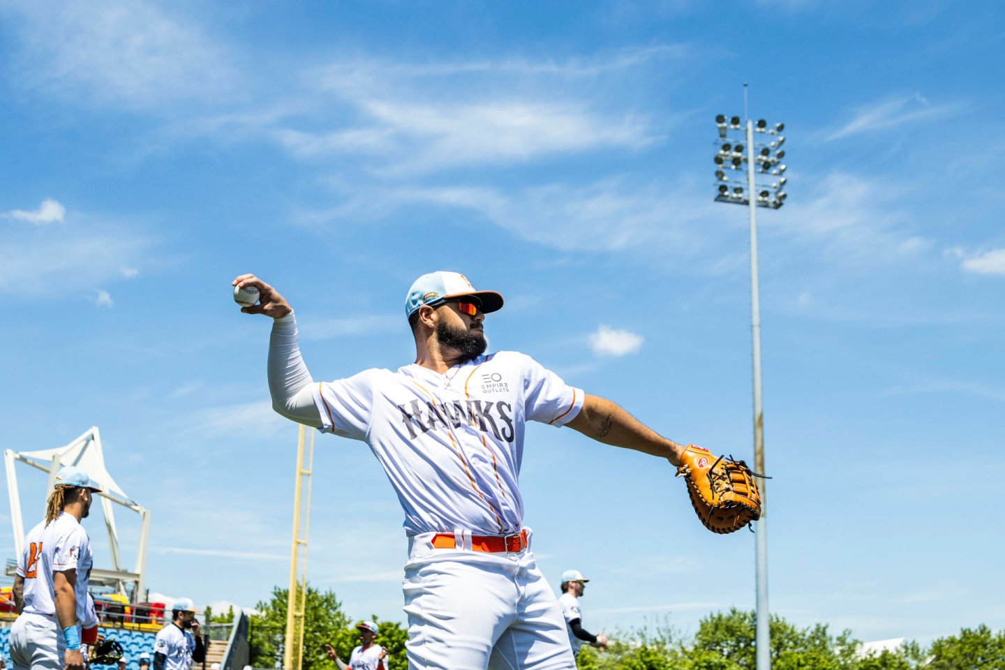 Baseball player in a white uniform and cap prepares to throw a ball on a sunny day. He holds a glove in his other hand. Teammates and trees are visible in the background under a clear blue sky.