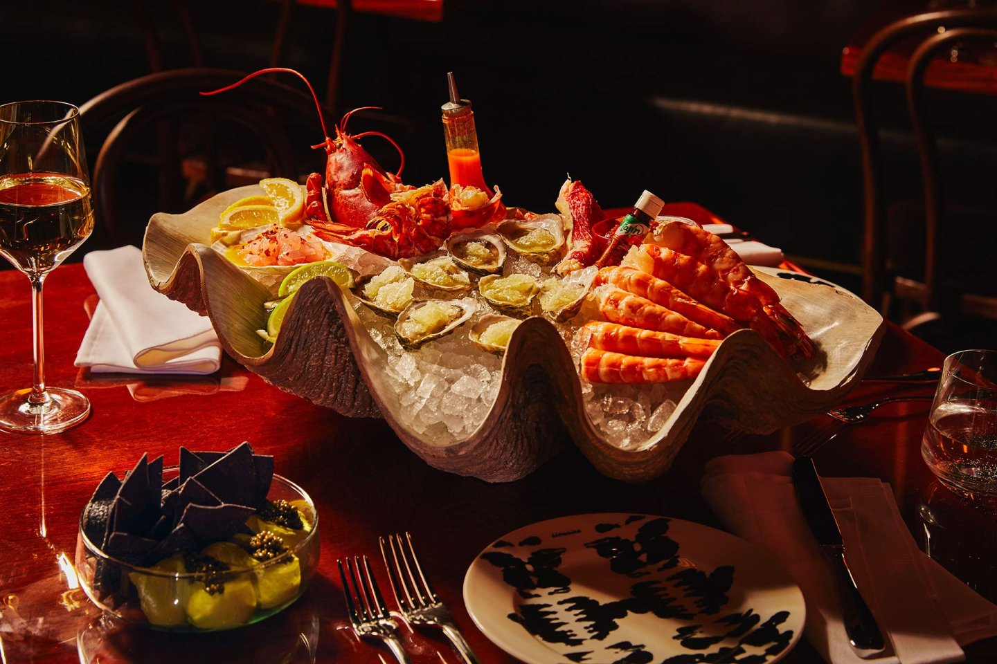 A large clam shell filled with oysters, prawns, lobster, and a small sauce bottle on ice sits on a red table, surrounded by wine glasses, forks, a plate, and a bowl of chips with guacamole.