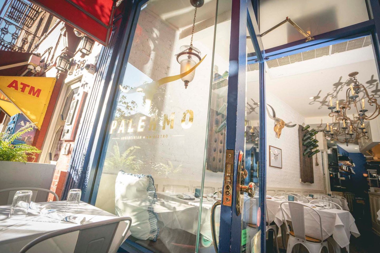 View of a restaurant entrance through glass doors with "Palermo" written on them. Inside, tables are set with white tablecloths and glassware. An ATM is visible outside under a yellow awning. The interior features a chandelier and wall decorations.