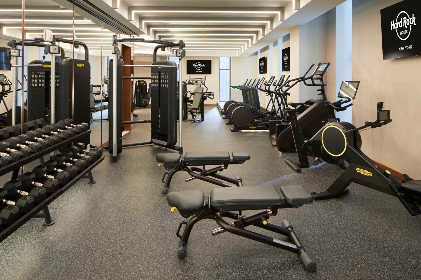 A spacious, modern gym with various exercise equipment including treadmills, exercise bikes, weight machines, dumbbells, and benches. The gym has bright lighting and a clean, organized layout with Hard Rock branding on the walls.