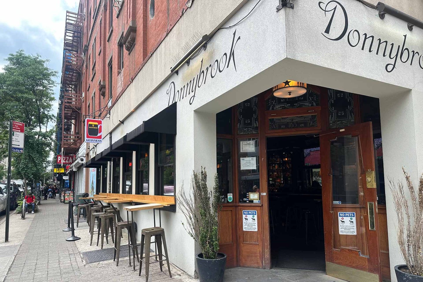 A street view of Donnybrook bar with open wooden doors, outdoor seating with stools along a counter, and a tree-lined sidewalk on a cloudy day. Signs and neighboring buildings are visible.