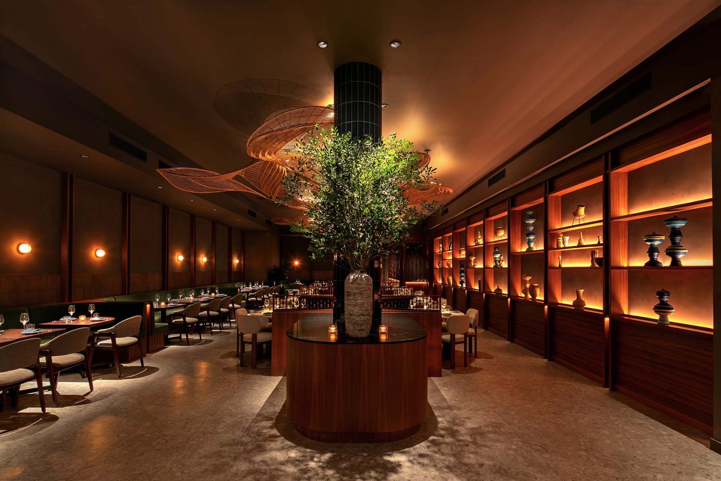 Elegant restaurant interior with warm lighting, wooden shelves displaying vases, neatly arranged tables and chairs, and a large potted plant as a centerpiece. The ambiance is cozy and inviting.