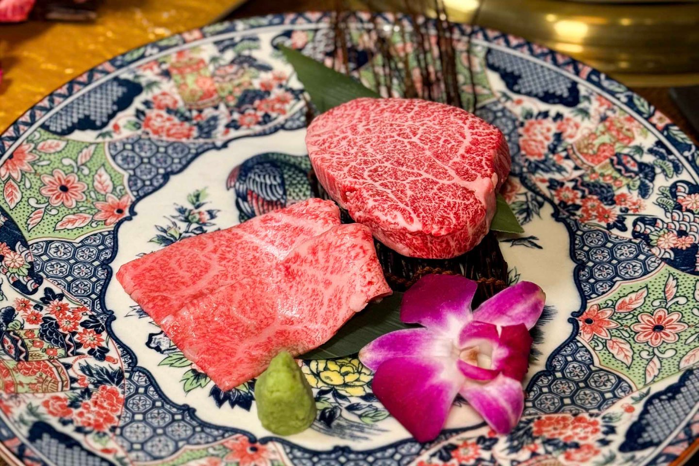 Two pieces of marbled raw beef, a wasabi dollop, and a purple orchid are arranged on a decorative floral plate with intricate blue and red designs.