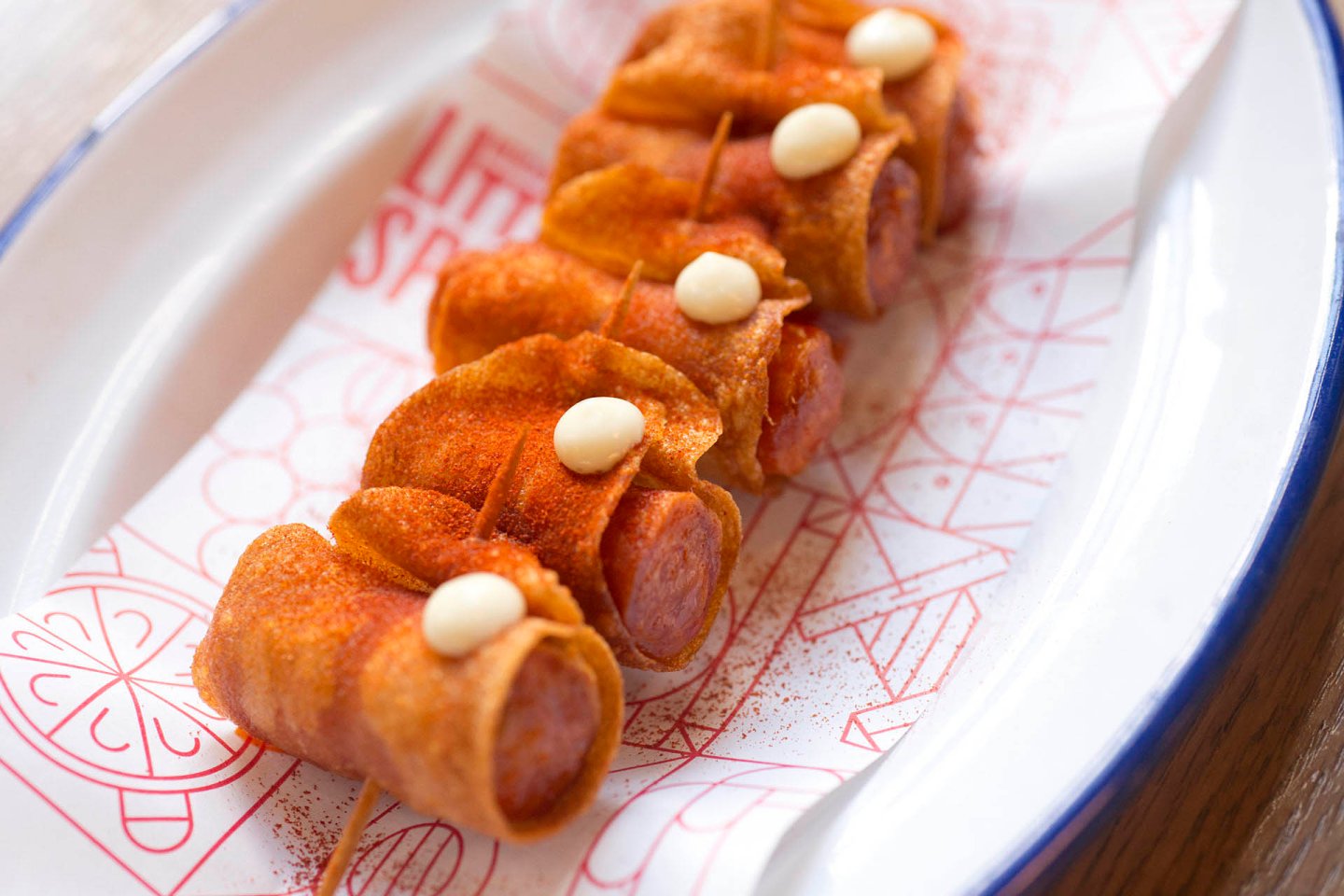 Skewered sausages wrapped in crispy, browned tortillas are served on a decorative red and white paper. The wraps are garnished with a light dusting of paprika and dollops of white sauce.