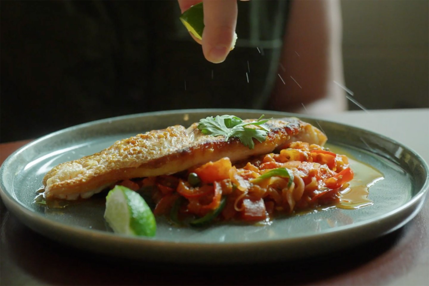 A hand squeezes a piece of lime over a plate of pan-seared fish served on a bed of sautéed vegetables, garnished with fresh herbs.