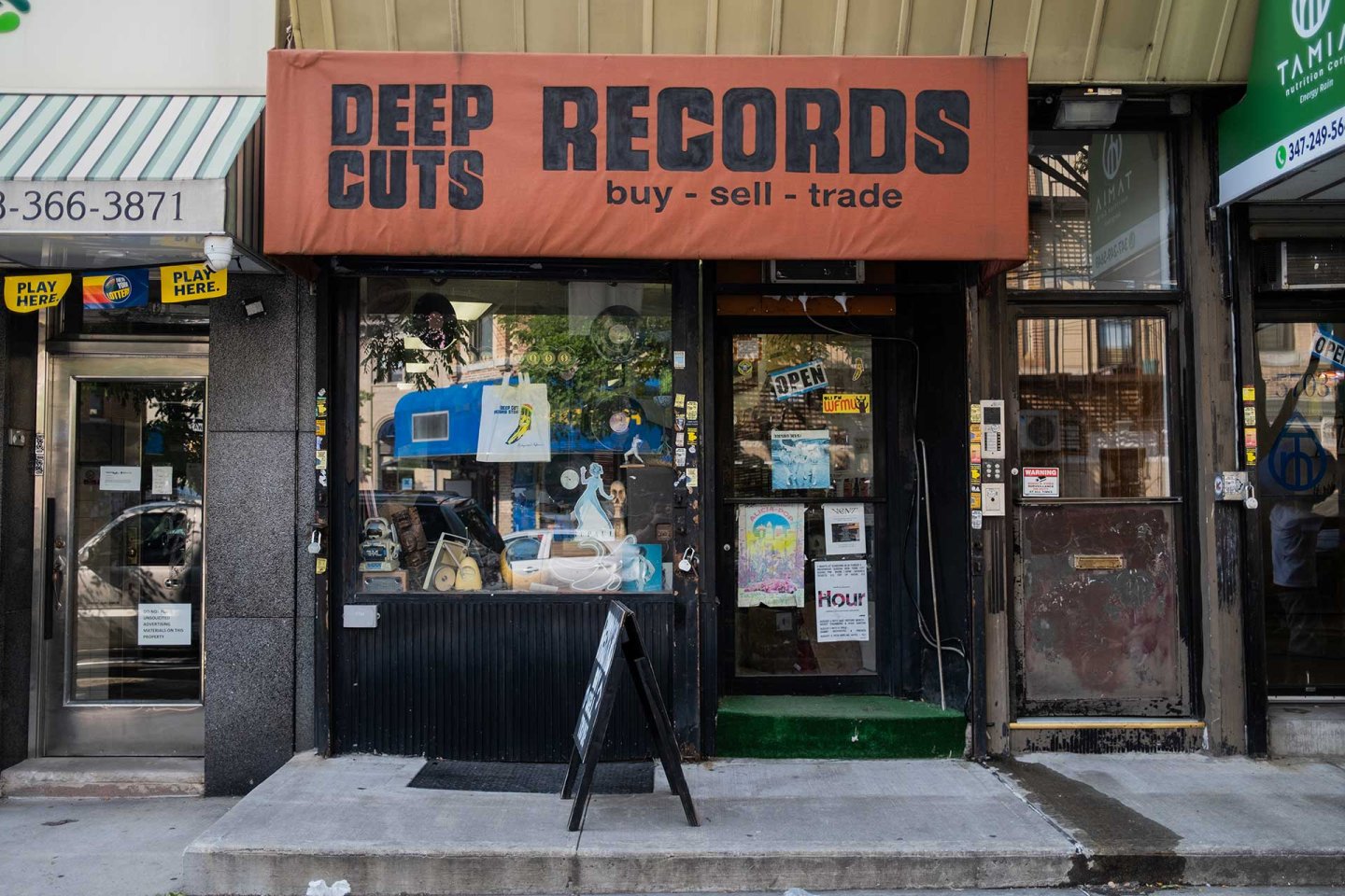 A storefront with an orange sign reading “DEEP CUTS RECORDS buy - sell - trade.” The window displays records and a neon “OPEN” sign. A sandwich board stands on the sidewalk.