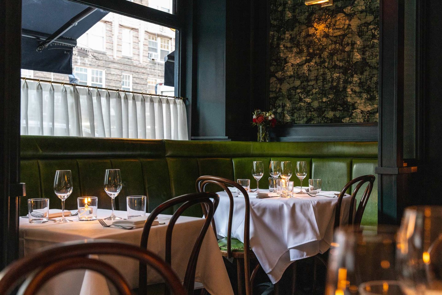Elegant restaurant interior with green leather seating, round tables covered in white tablecloths, set with wine glasses and candles. Large windows let in natural light, and a vase with flowers decorates the corner.