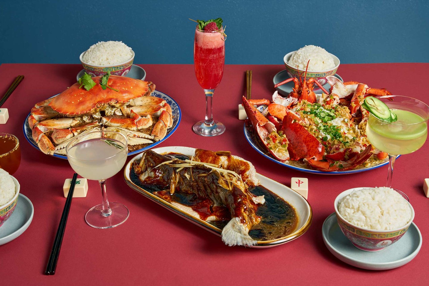 A table set with steamed crabs, lobster, a whole cooked fish with sauce, bowls of white rice, cocktails, and chopsticks, all arranged on red and blue tablecloths.