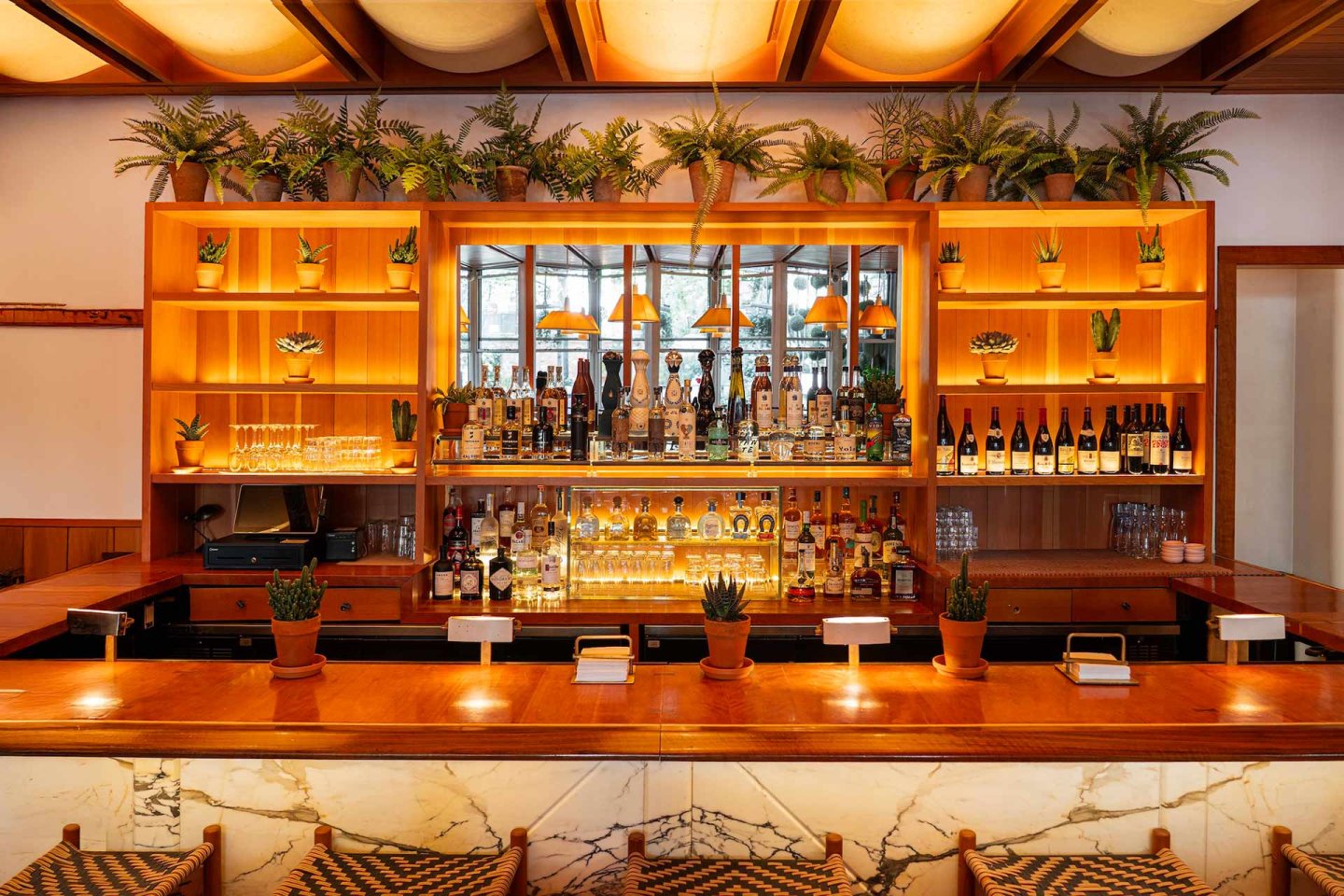A stylish bar with a wooden counter, shelves of liquor bottles, glassware, potted plants, and a mirrored back wall. Warm lighting creates a cozy atmosphere, and woven barstools line the counter.