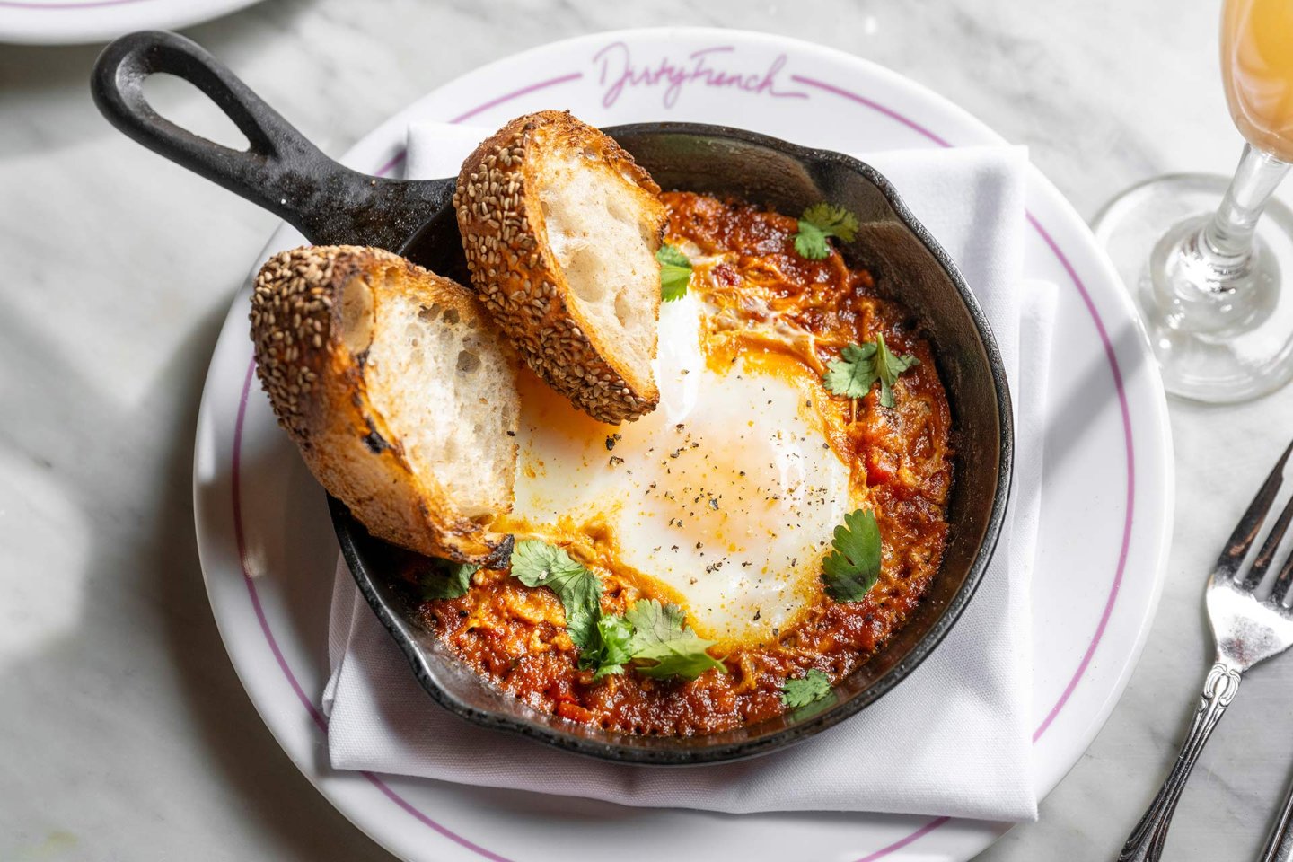 A cast-iron skillet with baked eggs in tomato sauce, garnished with fresh cilantro, served with two slices of seeded bread on a white plate. A fork and a glass of orange drink are nearby.
