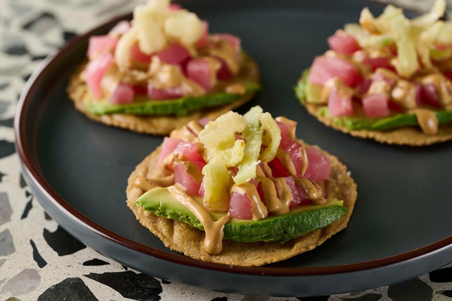 Three small tostadas on a dark plate, each topped with sliced avocado, diced raw tuna, creamy sauce, and crispy garnishes, set on a terrazzo surface.