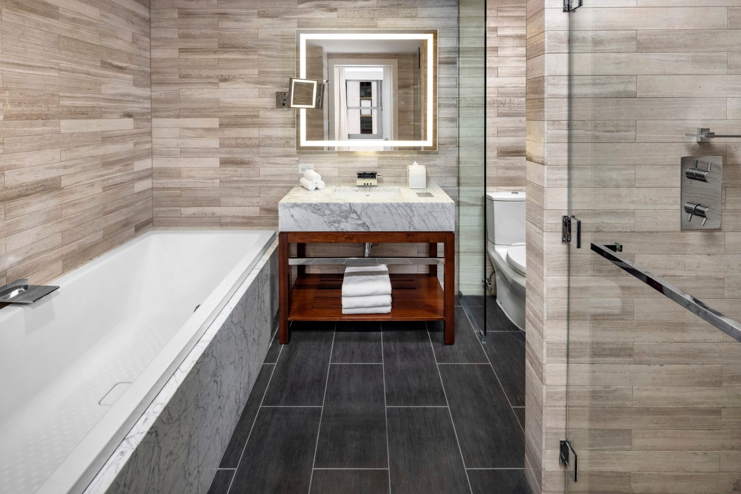 Modern bathroom with a marble bathtub, dark tiled floor, and a glass-enclosed shower. A wooden vanity with a marble countertop holds a mirror with backlighting. Towels are neatly stacked on a shelf under the sink. Walls are tiled in a light pattern.