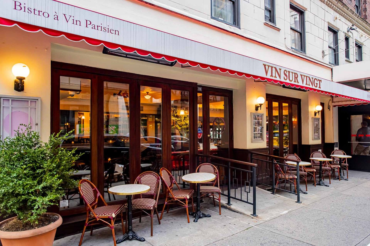 Outdoor view of a French bistro with a red and white awning, "Vin Sur Vingt," small round tables with wicker chairs, and large windows facing the street. A potted plant sits near the entrance.