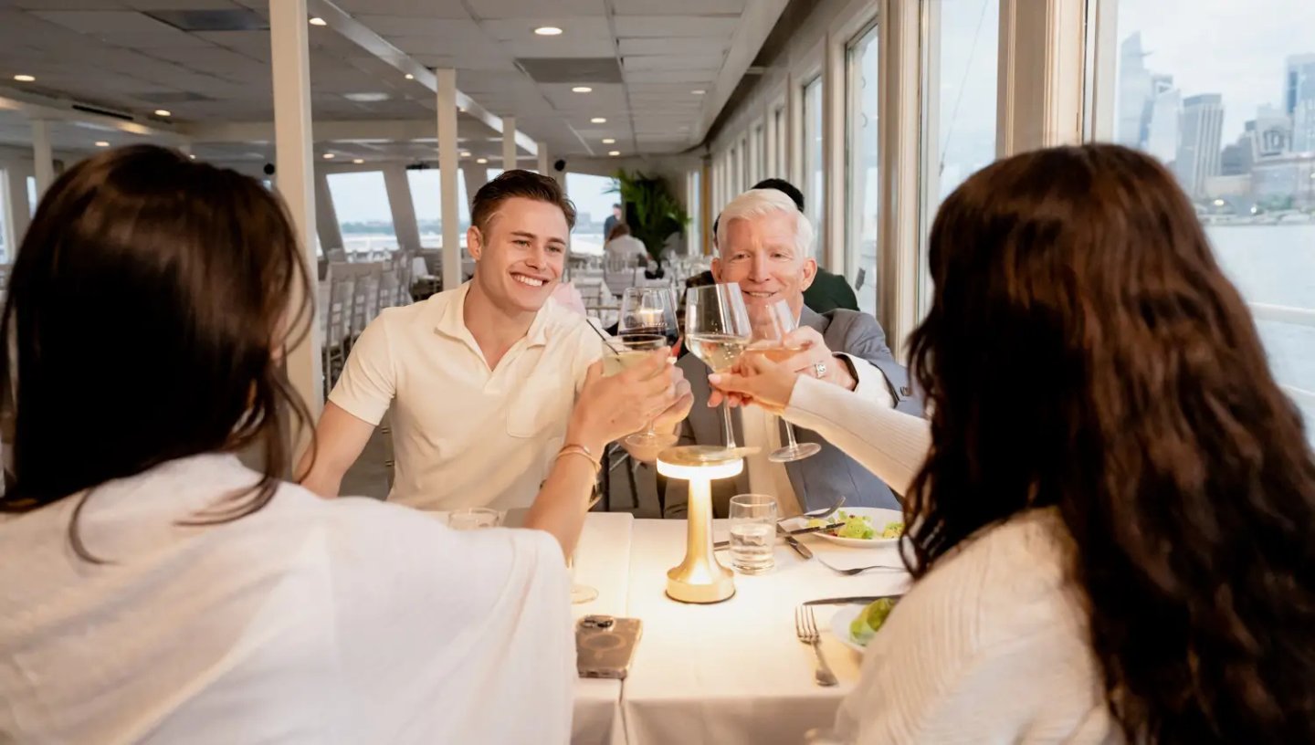 A smiling group of friends and family toast with wine and cocktail glasses while dining inside a well-lit boat cruise cabin with windows showing a city skyline view.