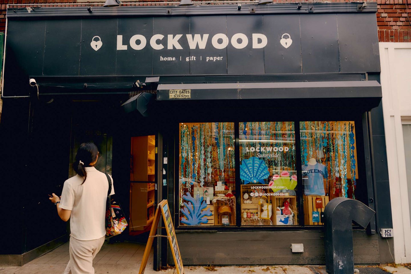 A person walks past the storefront of LOCKWOOD, a shop with a black sign reading "home | gift | paper." The window displays colorful decorations and various items for sale inside the store.