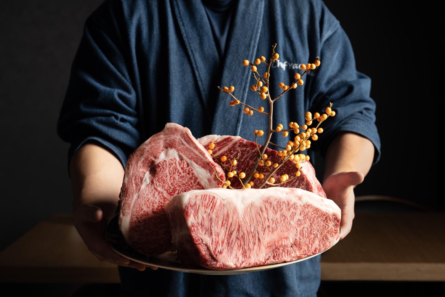 A person in a blue robe holds a large plate with three thick cuts of marbled raw beef, decorated with a sprig of orange berries.