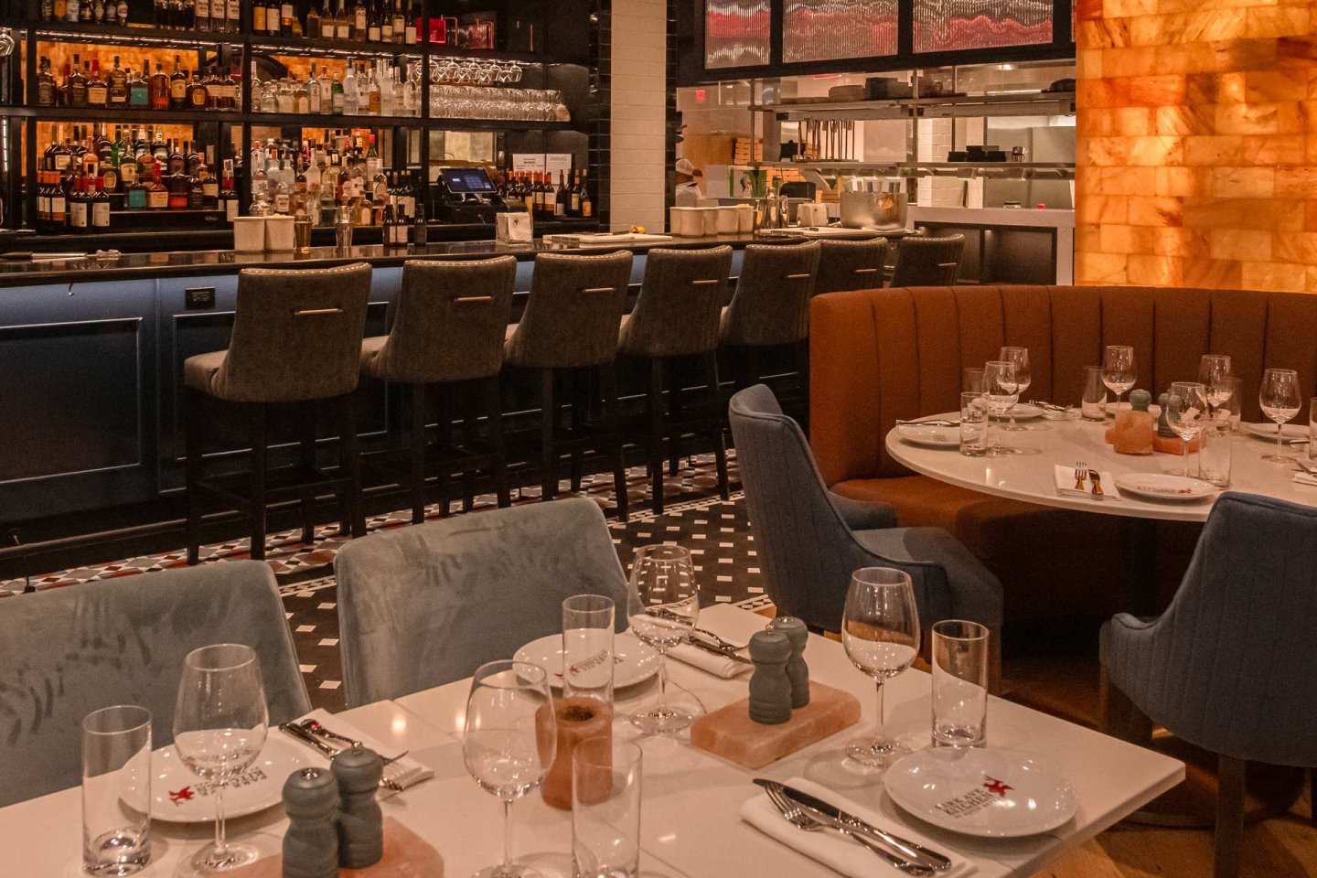 Cozy restaurant interior with elegantly set tables, cushioned chairs, and a bar lined with high-backed stools. Warm lighting highlights the decor, featuring neatly arranged glassware and tableware. Shelves behind the bar display an assortment of bottles.